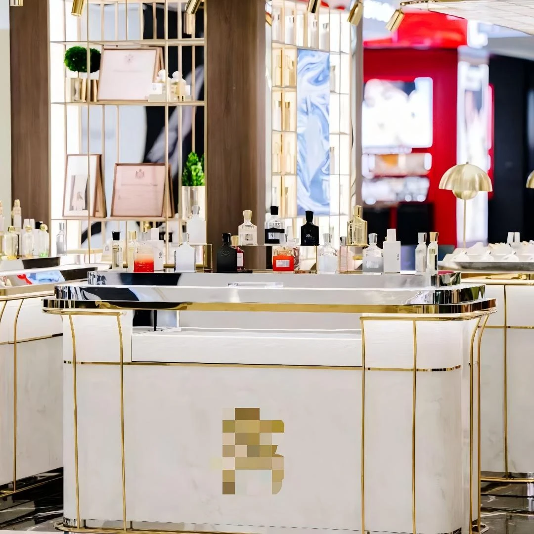 Perfume display case court under a gold canopy frame: curved white-marble counters with gold trims, lit gold shelving walls and velvet stools in a mall boutique