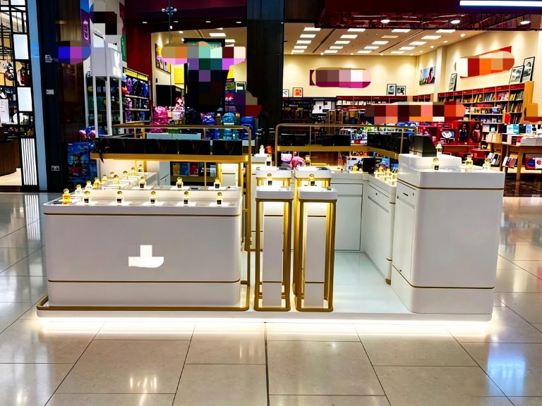 wide on-site view of the white and gold perfume kiosk showing tall gold-frame pedestals and rear shelving in the shopping centre